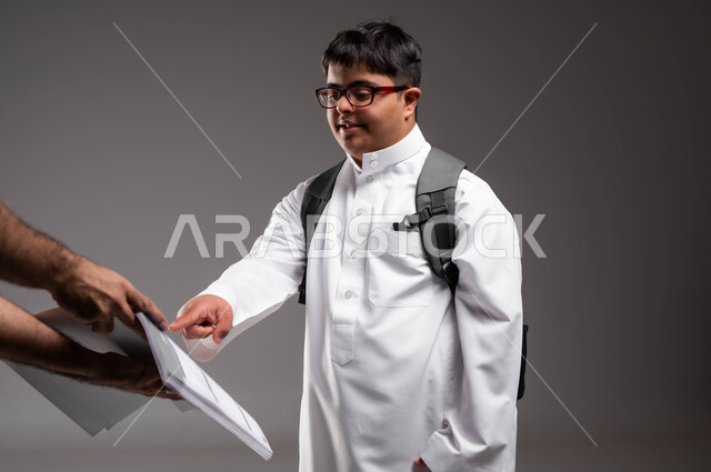 اختبارات و مذاكرات فصلية، الاشارة الى الأجوبة الصحيحة، بورتريه لشاب عربي خليجي سعودي مصاب بمتلازمة داون يرتدي الثوب التقليدي والنظارات الطبية و يحمل الحقيبة المدرسية، تطور وتقدم المناهج التعليمية، دمج ذوي الاحتياجات الخاصة في مدارس السعودية، خلفية رمادية .