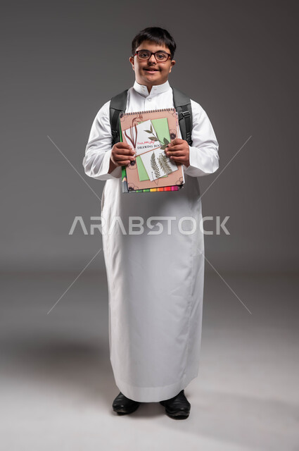 النظر الى الكاميرا بايماءات السرور، بورتريه لشاب عربي خليجي سعودي مصاب بمتلازمة داون يرتدي الثوب التقليدي والنظارات الطبية و يحمل الحقيبة و الكتب المدرسية، تطور وتقدم المناهج التعليمية، دمج ذوي الاحتياجات الخاصة في مدارس السعودية، صورة لكامل الجسم، خلفية رمادية .