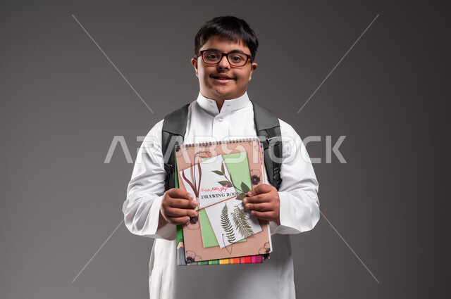 The development and progress of educational curricula, looking at the camera with happy gestures, a portrait of a smiling Gulf Arab Saudi man with Down Syndrome wearing a traditional dress and medical glasses and carrying a bag and textbooks, integrating people with special needs into Saudi schools, gray background.