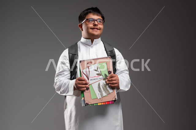 دمج ذوي الاحتياجات الخاصة في المدارس السعودية، النظر الى الاعلى لشيء ما، الإستعداد للعام الدراسي الجديد، بورتريه لشاب عربي خليجي سعودي مبتسم مصاب بمتلازمة داون يرتدي الثوب التقليدي والنظارات الطبية و يحمل الحقيبة و الكتب المدرسية، خلفية رمادية .
