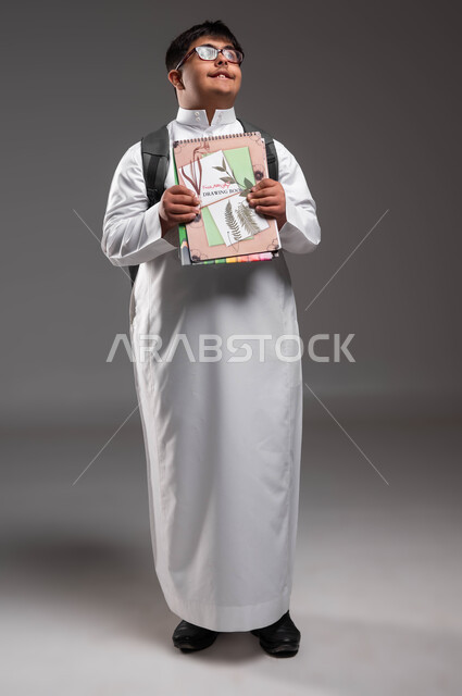 النظر الى الاعلى لشيء ما، الإستعداد للعام الدراسي الجديد، بورتريه لشاب عربي خليجي سعودي مبتسم مصاب بمتلازمة داون يرتدي الثوب التقليدي والنظارات الطبية و يحمل الحقيبة و الكتب المدرسية، دمج ذوي الاحتياجات الخاصة في المدارس السعودية، صورة لكامل الجسم، خلفية رمادية