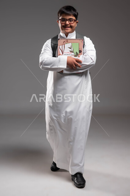 الإستعداد للعام الدراسي الجديد، النظر الى الكاميرا بايماءات السرور، بورتريه مقرب لشاب عربي خليجي سعودي مبتسم مصاب بمتلازمة داون يرتدي الثوب التقليدي والنظارات الطبية و يحمل الحقيبة و الكتب المدرسية، دمج ذوي الاحتياجات الخاصة في المدارس السعودية، خلفية رمادية