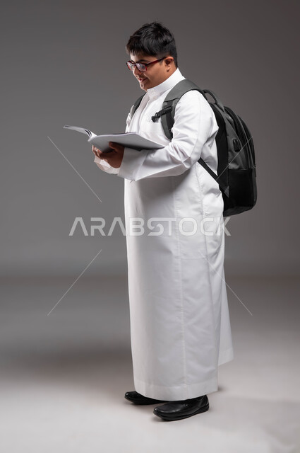 تطوير المناهج والمواد التعليمية، دمج ذوي الاحتياجات الخاصة في المدارس السعودية، بورتريه لطالب عربي خليجي سعودي يرتدي الثوب التقليدي وحقيبة الظهر المدرسية  مصاب بمتلازمة داون يحمل بيده الكتاب ويقوم بمراجعه الدروس ،صورة الجسم بالطول الكامل، خلفية رمادية .