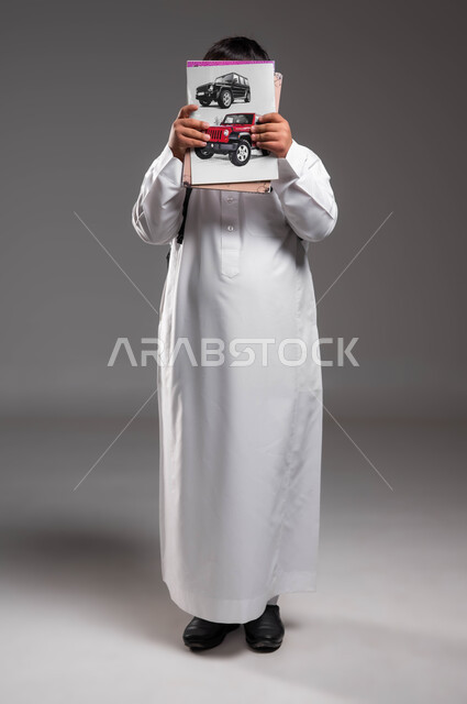 Displaying drawings of colorful SUVs, the concept of education in schools in the Kingdom of Saudi Arabia, a portrait of a Saudi Gulf Arab student wearing the traditional dress, holding books and school notebooks in his hands, practicing the hobby of drawing and coloring, full-length body portrait, gray background.