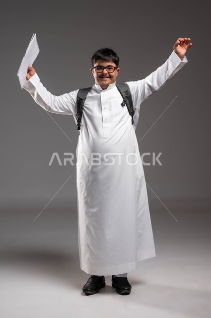 دمج ذوي الاحتياجات الخاصة في المدارس السعودية، رفع اليدين بايماءات السرور بالنجاح والتفوق، بورتريه لشاب عربي خليجي سعودي مبتسم مصاب بمتلازمة داون يرتدي الثوب التقليدي والنظارات الطبيه و يحمل الحقيبة المدرسية على ظهره، صورة الجسم بالطول الكامل، خلفية رمادية .