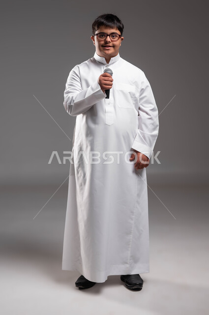 النظر الى الكاميرا بايماءات الثقة بالنفس، مشاركة ذوي الهمم في المسابقات الغنائية ،الاهتمام في المعاقين لشعورهم بالانتماء ، بورتريه لشاب عربي خليجي سعودي مصاب بمتلازمة داون يرتدي الزي التقليدي ويحمل بيده المايكروفون ،صورة لكامل الجسم، خلفية رمادية .