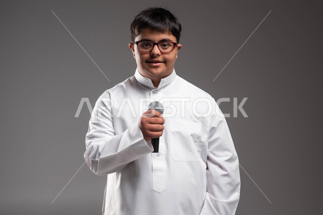 Standing straight and looking at the camera, participation of people of determination in singing competitions, caring for the disabled for their sense of belonging and appreciation, a close-up portrait of a young Saudi Arabian Gulf man with Down Syndrome, wearing traditional clothing and medical glasses and holding a microphone in his hand, gray background.