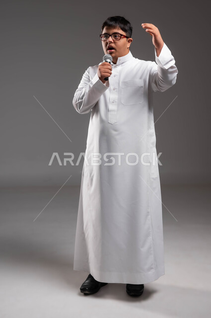 ايماءات الاندماج اثناء تقديم العروض الفنيه الغنائيه، انشطة ترفيهيه ممتعه، مشاركة ذوي الهمم في المجتمع السعودي، بورتريه لشاب عربي خليجي سعودي مصاب بمتلازمة داون يرتدي الزي التقليدي و النظارات الطبية ويحمل بيده المايكروفون، صورة لكامل الجسم،خلفية رمادية .