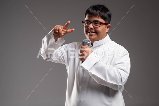 رفع اليد بتعابير و اشارات مختلفة ،مشاركة ذوي الهمم في المسابقات الغنائية ،الاهتمام في المعاقين لشعورهم بالانتماء والتقدير، بورتريه مقرب لشاب عربي خليجي سعودي مبتسم مصاب بمتلازمة داون يرتدي الزي التقليدي و النظارات الطبيه ويحمل بيده المايكروفون، خلفية رمادية .