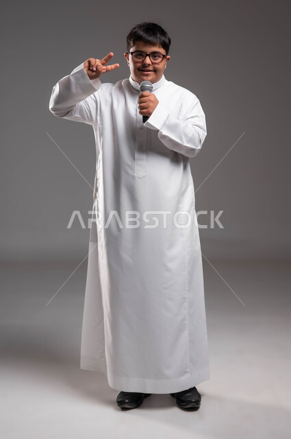 رفع اليد باشارات وتعابير مختلفة، مشاركة ذوي الهمم في المسابقات الغنائية ،الاهتمام في المعاقين لشعورهم بالحب والتقدير، بورتريه لشاب عربي خليجي سعودي مصاب بمتلازمة داون يرتدي الزي التقليدي و النظارات الطبيه ويحمل بيده المايكروفون، صورة لكامل الجسم، خلفية رمادية .