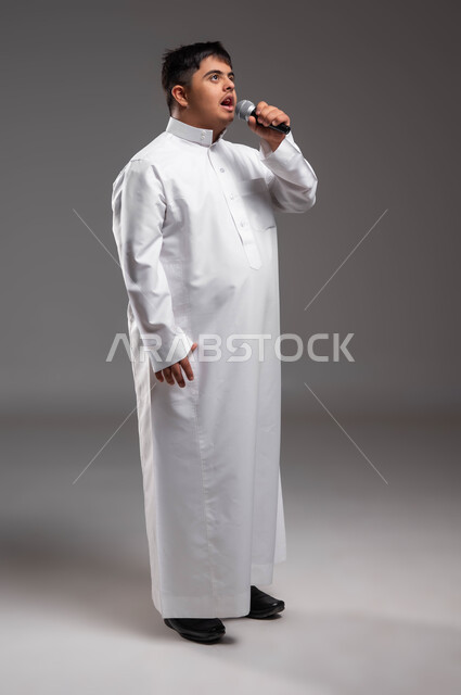 مشاركة ذوي الهمم في المسابقات الغنائية ،الاهتمام في المعاقين لشعورهم بالانتماء و التقدير، بورتريه لشاب عربي خليجي سعودي مصاب بمتلازمة داون يرتدي الزي التقليدي ويحمل بيده المايكروفون ويقوم بالغناء، تقديم العروض الفنيه المسرحيه، صورة لكامل الجسم، خلفية رمادية .
