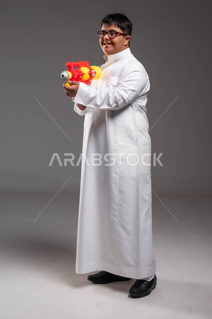 النظر الى شيء ما بإيماءات السرور، انشطة ترفيهيه مسلية، قضاء اوقات ممتعه بالألعاب المائية في فصل الصيف، بورتريه لشاب عربي خليجي سعودي مصاب بمتلازمة داون يرتدي الزي التقليدي و النظارات الطبية يحمل بيده مسدس مائي بلاستيكي، صورة للجسم بالكامل، خلفية رمادية .