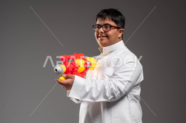 The concept of self-recreation and amusement, spending enjoyable times with water games in the summer, a close-up portrait of a young Saudi Arabian Gulf man with Down Syndrome, wearing traditional clothing and medical glasses, holding a plastic water gun in his hand and looking at something with happy gestures, gray background.
