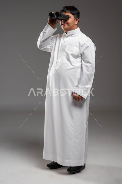 The sense of belonging and appreciation for people with disabilities in Saudi Arabia, the love of exploration and adventure, a future vision for people with disabilities, a portrait of a Saudi Gulf Arab young man wearing traditional clothing with Down Syndrome, holding binoculars in his hand, full-length body photo, gray background