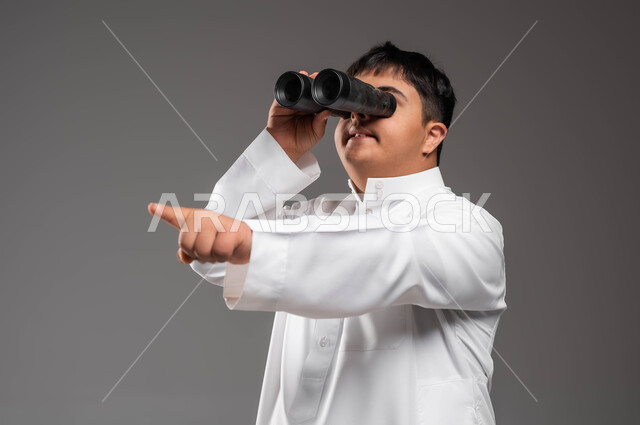 رؤية مستقبلية عند أصحاب ذوي الهمم، بورتريه مقرب لشاب عربي خليجي سعودي يرتدي الزي التقليدي مصاب بمتلازمة داون يحمل بيده المنظار ثنائي العينين ويشير الى شيء ما، شعور المعاقين بالإنتماء والتقدير في السعوديه، حب الاستكشاف والمغامرة ،خلفية رمادية