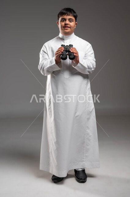 Seeing distant things through binoculars, future planning for goals and ambitions among people of determination, portrait of a smiling Saudi Arabian Gulf man with Down Syndrome wearing a traditional dress holding binoculars in his hand, full body image, love of exploration and adventure, gray background