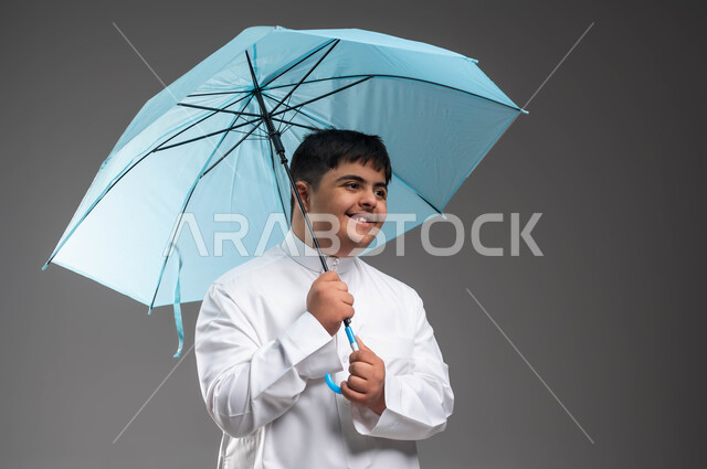 التظلل من أشعة الشمس، تعابير وايماءات الحماس والسرور، بورتريه مقرب لشاب عربي خليجي سعودي مبتسم مصاب بمتلازمة داون يرتدي الثوب التقليدي يحمل بيده مظلة زرقاء، الحماية من الأمطار في الشتاء، دمج ذوي الهمم بالمجتمع السعودي، خلفية رمادية