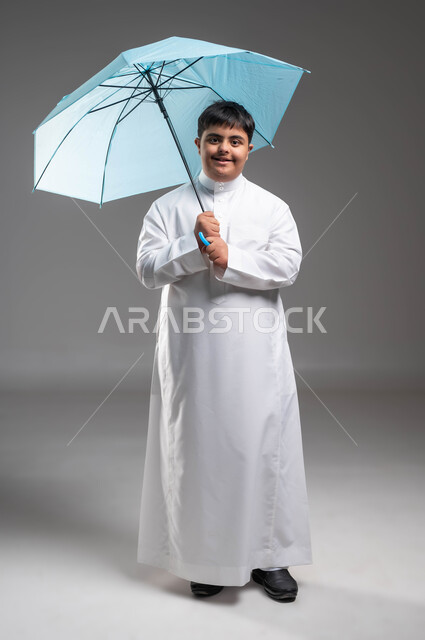 الحماية من الأمطار في الشتاء، النظر الى الكاميرا بتعابير السرور والسعادة، بورتريه لشاب عربي خليجي سعودي مبتسم مصاب بمتلازمة داون يرتدي الثوب التقليدي يحمل بيده مظلة زرقاء، دمج ذوي الهمم بالمجتمع السعودي، التظلل من أشعة الشمس، صورة الجسم بالكامل، خلفية رمادية
