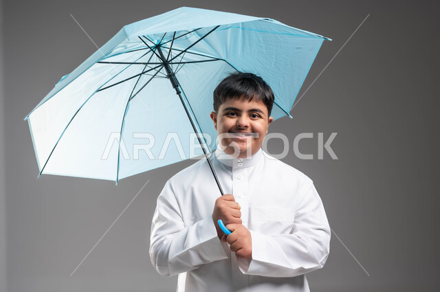 النظر الى الكاميرا بتعابير وايماءات السرور والسعادة، بورتريه مقرب لشاب عربي خليجي سعودي مبتسم مصاب بمتلازمة داون يرتدي الثوب التقليدي يحمل بيده مظلة زرقاء، الحماية من الأمطار في الشتاء، دمج ذوي الهمم بالمجتمع السعودي، التظلل من أشعة الشمس، خلفية رمادية
