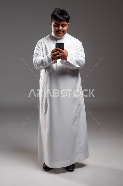 تعليم المعاقين على استخدام الأجهزة التقنية المتطورة، بورتريه لشاب عربي خليجي سعودي مبتسم مصاب بمتلازمة داون يرتدي الثوب التقليدي يستخدم الهاتف المحمول، تعابير وايماءات الاندماج والسرور، صورة الجسم بالطول الكامل، خلفية رمادية