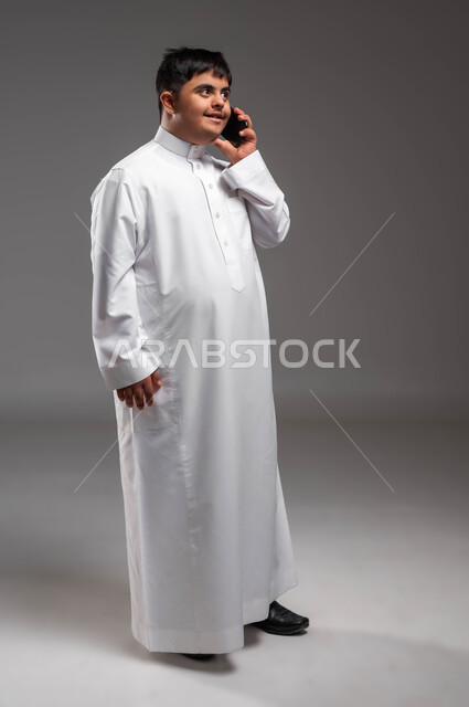 الدردشة والتواصل مع الأصدقاء والاقارب، تعليم المعاقين على استخدام الاجهزة التقنية الحديثة، بورتريه لشاب عربي خليجي سعودي مبتسم مصاب بمتلازمة داون يرتدي الثوب التقليدي يقوم بإجراء مكالمة هاتفية عبر الجوال بتعابير السرور، صورة لكامل الجسم، خلفية رمادية