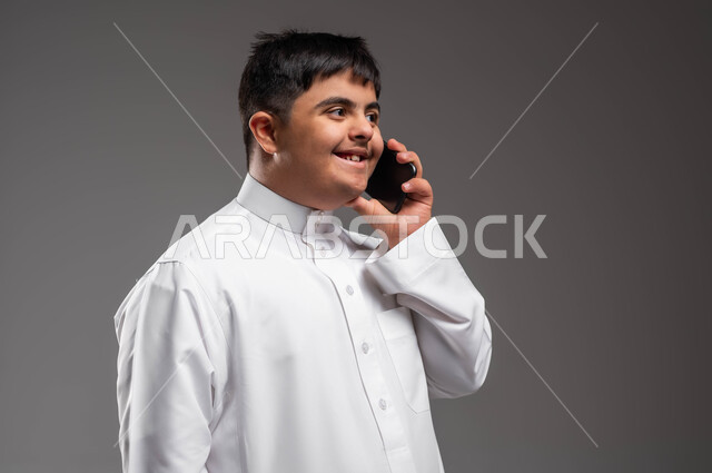 التواصل والدردشة مع الأصدقاء والاقارب، تعليم المعاقين على استخدام الاجهزة التقنية الحديثة، بورتريه مقرب لشاب عربي خليجي سعودي مبتسم مصاب بمتلازمة داون يرتدي الثوب التقليدي يقوم بإجراء مكالمة هاتفية عبر الجوال، قضاء وقت ممتع بتبادل الأحاديث، خلفية رمادية