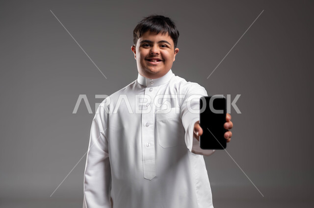 عرض تطبيقات لأصحاب الاحتياجات الخاصة عبر الهاتف المحمول، بورتريه مقرب لشاب عربي خليجي سعودي مبتسم مصاب بمتلازمة داون يرتدي ثوب تقليدي يحمل بيده جوال بشاشة سوداء فارغة، تسهيل حياة ذوي الهمم باستخدام التكنولوجيا، تعليم المعاقين على الأجهزة التقنية, خلفية رمادية