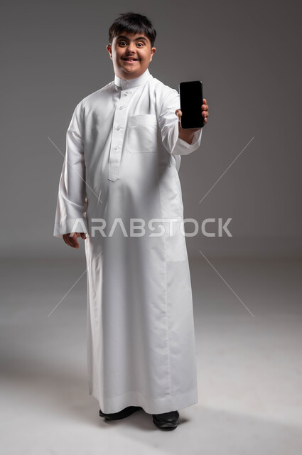 Facilitating the lives of people of determination using technology and technical devices, offering applications for people with special needs via mobile phones, a portrait of a smiling Saudi Arabian Gulf man with Down Syndrome, wearing a traditional dress, holding a mobile phone with a blank black screen in his hand, a full-body photo, gray background.