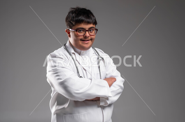 دمج ذوي الإعاقة في الحياه المهنيه والعمليه, بورتريه مقرب لطبيب عربي خليجي سعودي مبتسم مصاب بمتلازمة داون يرتدي المعطف الابيض والسماعة الطبية يقف بأيدي متقاطعة، الفحص الطبي وتقديم الخدمات الصحية للمرضى، تمكين أصحاب الهمم من الحصول على فرص عمل، خلفية رمادية