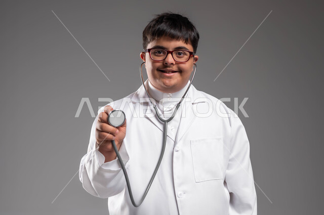 دمج ذوي الإعاقة في الحياه المهنيه والعمليه, بورتريه مقرب لطبيب عربي خليجي سعودي مبتسم مصاب بمتلازمة داون يرتدي المعطف الابيض والسماعة الطبية، الفحص الطبي وتقديم الخدمات الصحية للمرضى، تمكين أصحاب الهمم من الحصول على فرص عمل، خلفية رمادية