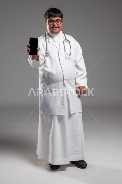استخدام الأجهزة الالكترونية في المجال الطبي، دمج ذوي الإعاقة في الحياه المهنيه, بورتريه لطبيب عربي خليجي سعودي مصاب بمتلازمة داون يرتدي المعطف الابيض والسماعة الطبية يقوم بعرض جوال بشاشة سوداء فارغة، توفير فرص عمل لأصحاب الهمم، صورة لكامل الجسم، خلفية رمادية