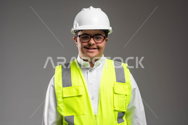 Enabling people of determination to obtain suitable job opportunities, integrating people with disabilities into professional and practical life, close-up portrait of a smiling Saudi Arabian Gulf engineer with Down Syndrome wearing a helmet, protection vest, and medical glasses, looking at something with an expression of pleasure, gray background.