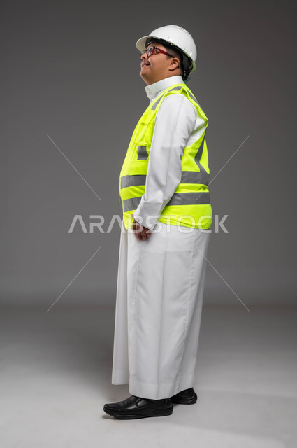 دمج المعاقين في الأعمال والمهن السعودية,  تمكين أصحاب الهمم من الحصول على فرص عمل مناسبة, بورتريه من الجانب لمهندس عربي خليجي سعودي مبتسم مصاب بمتلازمة داون يرتدي الخوذه وسترة الحماية والنظارة الطبية ينظر إلى لشيء ما بتعابير السرور، صورة لكامل الجسم، خلفية رمادية