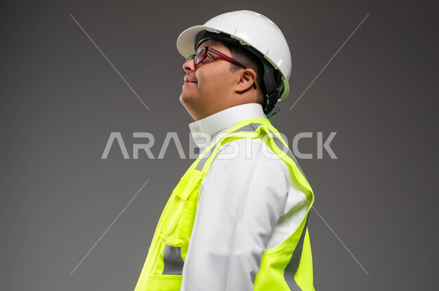 تمكين أصحاب الهمم من الحصول على فرص عمل مناسبة, دمج ذوي الإعاقة في الحياه المهنيه والعمليه, بورتريه مقرب من الجانب لمهندس عربي خليجي سعودي مبتسم مصاب بمتلازمة داون يرتدي الخوذه وسترة الحماية والنظارة الطبية ينظر شيء إلى ما بتعابير السرور، خلفية رمادية