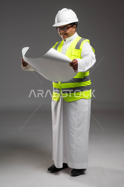 متابعة الأعمال الهندسية، دمج المعاقين في المهن السعودية,  تمكين أصحاب الهمم من الحصول على فرص عمل مناسبة, بورتريه لمهندس عربي خليجي سعودي مبتسم مصاب بمتلازمة داون يرتدي الخوذه وسترة الحماية والنظارة الطبية يقوم بتدقيق مخطط البناء، صورة لكامل الجسم، خلفية رمادية