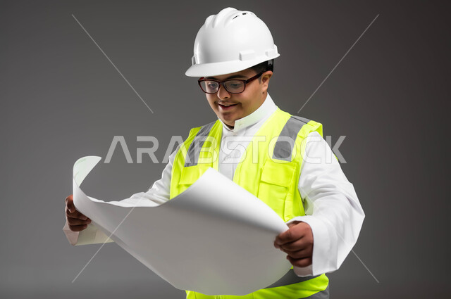 مشاركة ذوي الاحتياجات بالأعمال الهندسية، تمكين أصحاب الهمم من الحصول على فرص عمل مناسبة, بورتريه مقرب لمهندس عربي خليجي سعودي مبتسم مصاب بمتلازمة داون يرتدي الخوذه وسترة الحماية والنظارة الطبية يقوم بتدقيق مخطط البناء، خلفية رمادية