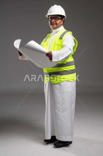 متابعة الأعمال الهندسية، دمج المعاقين في المهن السعودية,  تمكين أصحاب الهمم من الحصول على فرص عمل مناسبة, بورتريه لمهندس عربي خليجي سعودي مبتسم مصاب بمتلازمة داون يرتدي الخوذه وسترة الحماية والنظارة الطبية يقوم بتدقيق مخطط البناء، صورة لكامل الجسم، خلفية رمادية