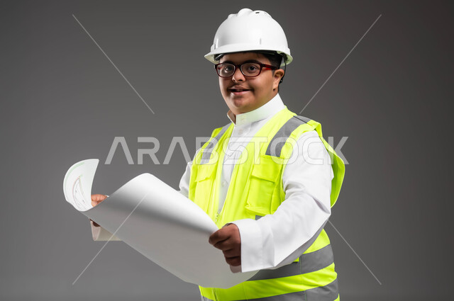 مشاركة ذوي الاحتياجات بالأعمال الهندسية، تمكين أصحاب الهمم من الحصول على فرص عمل مناسبة, بورتريه مقرب لمهندس عربي خليجي سعودي مبتسم مصاب بمتلازمة داون يرتدي الخوذه وسترة الحماية والنظارة الطبية يقوم بتدقيق مخطط البناء، خلفية رمادية