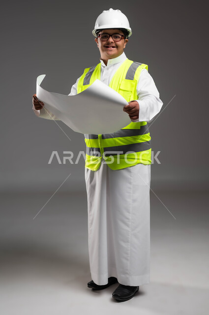 استمتاع المعاقين بالمشاركة بالمهن والأعمال الهندسية, تمكين أصحاب الهمم من الحصول على فرص عمل مناسبة, بورتريه لمهندس عربي خليجي سعودي مبتسم مصاب بمتلازمة داون يرتدي الخوذه وسترة الحماية والنظارة الطبية يحمل مخطط البناء، صورة لكامل الجسم، خلفية رمادية