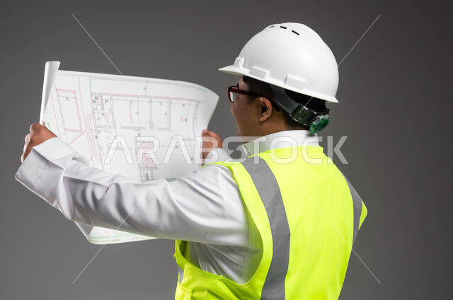 Supervising engineering works, enabling people of determination to obtain suitable job opportunities, close-up portrait of a smiling Saudi Arabian Gulf engineer with Down Syndrome wearing a helmet, protection vest, and medical glasses, checking the building plan, gray background
