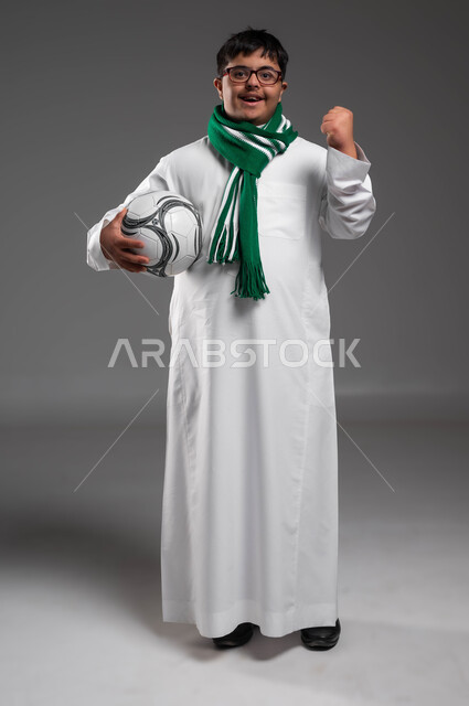 تنمية الهوايات الرياضية لذوي الإعاقة, النظر إلى الكاميرا بتعابير التشجيع والفوز، ارتداء أوشحة تشجيعية للفريق المفضل, بورتريه لشاب عربي خليجي سعودي مبتسم مصاب بمتلازمة داون يرتدي النظارة الطبية يحمل بيده كرة القدم، صورة الجسم بالكامل، خلفية رمادية