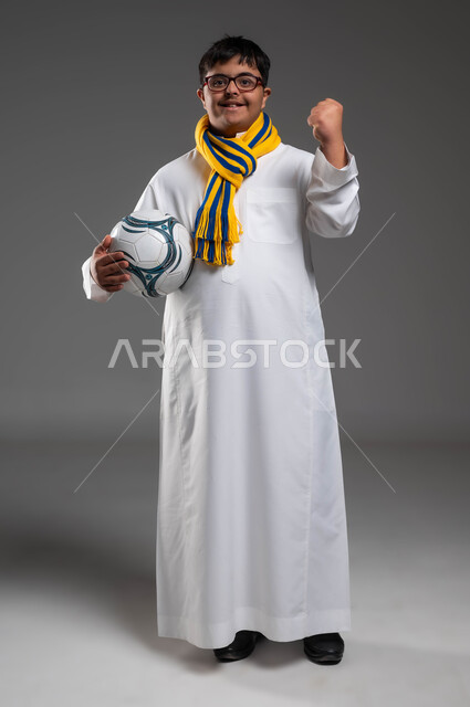 تنمية الهوايات الرياضية لذوي الإعاقة, النظر إلى الكاميرا بتعابير التشجيع والفوز، ارتداء أوشحة تشجيعية للفريق المفضل, بورتريه لشاب عربي خليجي سعودي مبتسم مصاب بمتلازمة داون يرتدي النظارة الطبية يحمل بيده كرة القدم، صورة الجسم بالكامل، خلفية رمادية