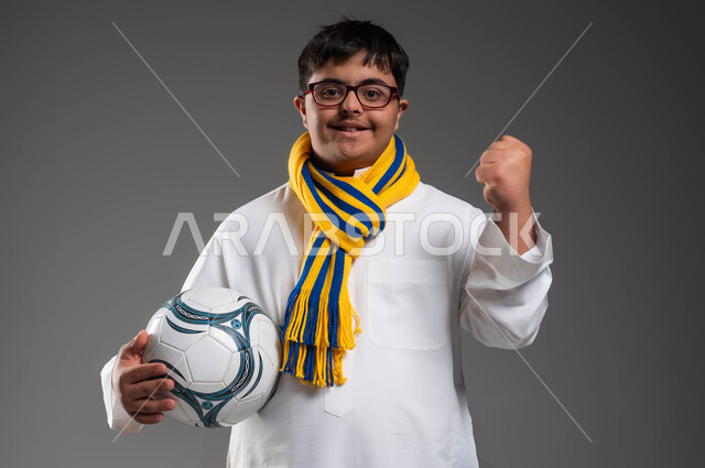Wearing scarves for the favorite team, looking at the camera with expressions of encouragement and victory, developing sports hobbies for people with disabilities, close-up portrait of a smiling Saudi Gulf Arab young man with Down Syndrome, wearing glasses, holding a football in his hand, gray background