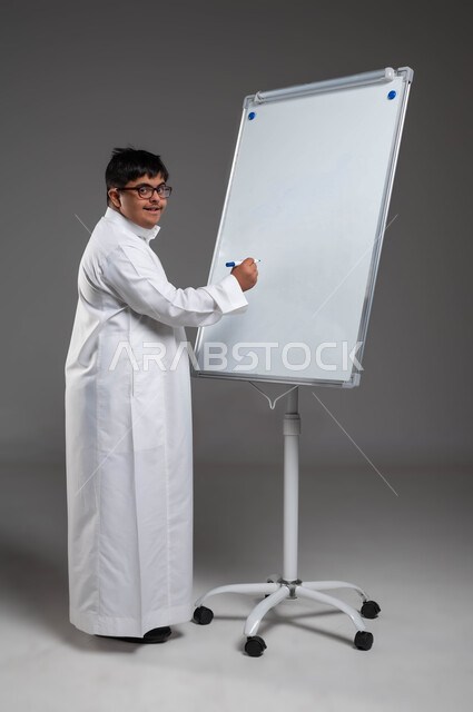 اهتمام المجتمع السعودي بتعليم ذوي الهمم, تنمية القدرات الفنية والفكرية للمعاقين, بورتريه لشاب عربي خليجي سعودي مبتسم مصاب بمتلازمة داون يرتدي الثوب التقليدي والنظارة الطبية يكتب على بورد أبيض, القيام بالأنشطة الترفيهية والتثقيفية، صورة لكامل الجسم، خلفية رمادية