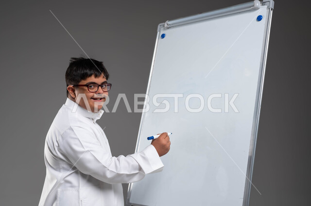 تنمية القدرات الفنية والفكرية للمعاقين, اهتمام المجتمع السعودي بتعليم ذوي الهمم, بورتريه مقرب لشاب عربي خليجي سعودي مبتسم مصاب بمتلازمة داون يرتدي الثوب التقليدي والنظارة الطبية يقوم بالكتابة على بورد أبيض, القيام بالأنشطة الترفيهية والتثقيفية، خلفية رمادية