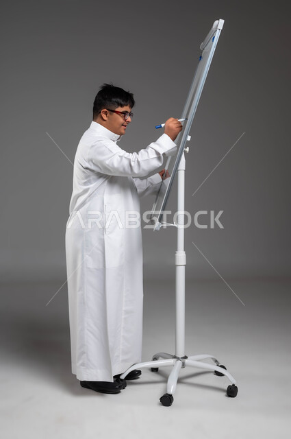 اهتمام المجتمع السعودي بتعليم ذوي الهمم, تنمية القدرات الفنية والفكرية للمعاقين, بورتريه لشاب عربي خليجي سعودي مصاب بمتلازمة داون يرتدي الثوب التقليدي والنظارة الطبية يكتب على بورد أبيض, القيام بالأنشطة الترفيهية والتثقيفية، صورة لكامل الجسم، خلفية رمادية