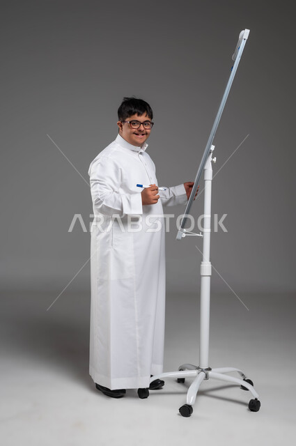 Developing the artistic and intellectual capabilities of people with disabilities, Saudi society’s interest in educating people of determination, a portrait of a smiling Saudi Gulf Arab man with Down Syndrome, wearing a traditional dress and medical glasses, writing on a white board, doing recreational and educational activities, a full-body portrait, gray background