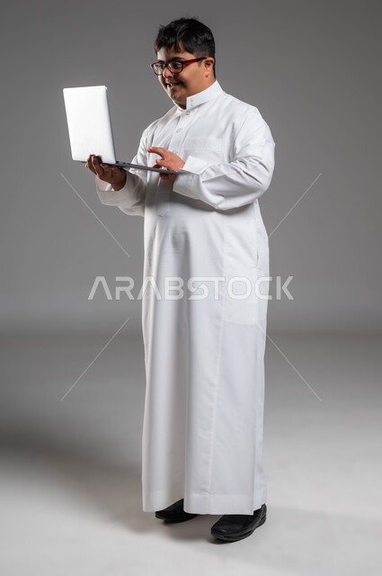 محاولة المجتمع السعودي تنمية قدرات أصحاب الهمم, دمج ذوي الاحتياجات الخاصه في الحياه المهنيه والعمليه, بورتريه لشاب عربي خليجي سعودي مبتسم مصاب بمتلازمة داون يرتدي الثوب التقليدي والنظارة الطبية يستخدم الحاسب المحمول, صورة الجسم بالكامل، خلفية رمادية