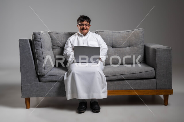 النظر إلى الكاميرا بابتسامة، دمج ذوي الاحتياجات الخاصه في الحياه المهنيه والعمليه, بورتريه لشاب عربي خليجي سعودي مبتسم مصاب بمتلازمة داون يرتدي الثوب التقليدي والنظارة الطبية يستخدم الحاسب المحمول, اهتمام المجتمع السعودي بقدرات أصحاب الهمم, خلفية رمادية