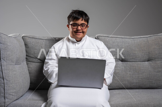 النظر إلى الكاميرا بابتسامة، دمج ذوي الاحتياجات الخاصه في الحياه المهنيه والعمليه, بورتريه لشاب عربي خليجي سعودي مبتسم مصاب بمتلازمة داون يرتدي الثوب التقليدي والنظارة الطبية يستخدم الحاسب المحمول, اهتمام المجتمع السعودي بقدرات أصحاب الهمم, خلفية رمادية